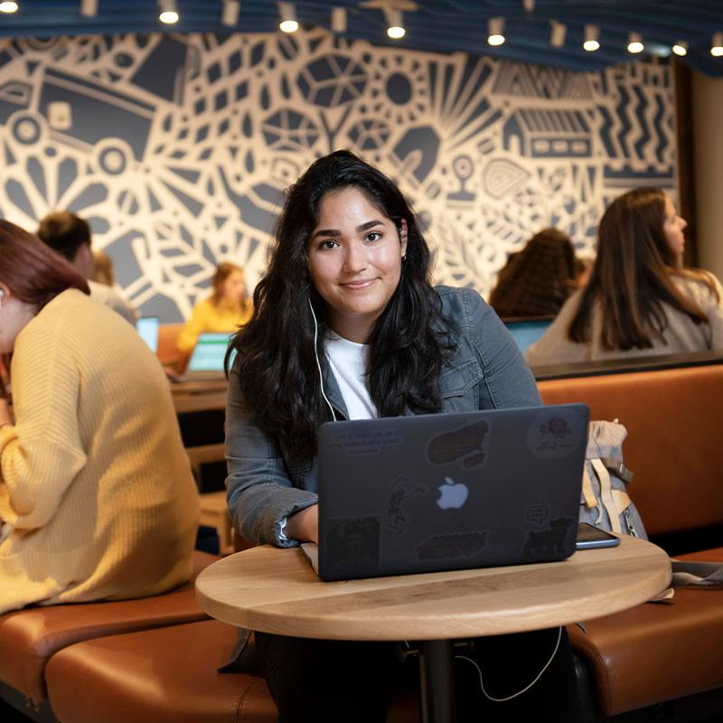 A Drake University student smiling while working on a laptop sitting at a single table in a dining location on campus